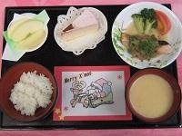 赤いお盆に置かれたご飯、魚の切り身と付け合わせの野菜や、スープ、りんご、ケーキとトナカイとサンタのイラストが描かれたカードの写真
